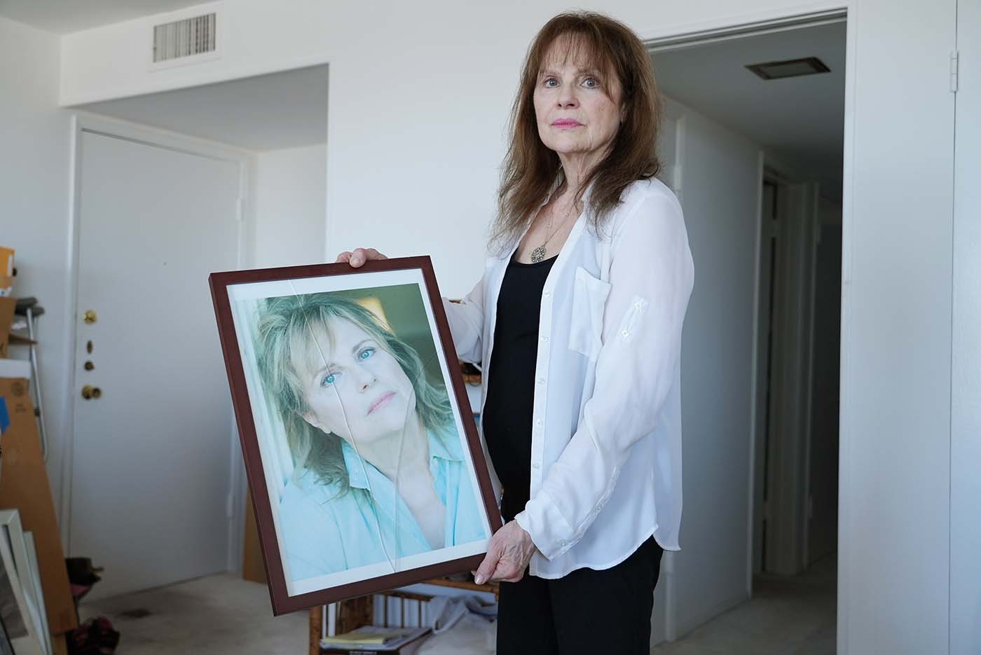 Janice Lynde holds stands in her apartment holding a broken framed photo.